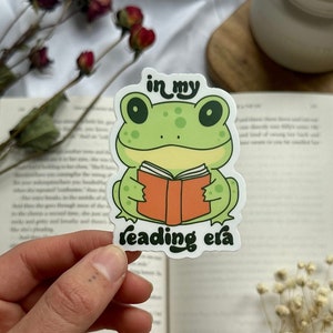 Könnte beinhalten: Ein grüner Frosch-Aufkleber mit einem Buch in seinen Händen. Der Aufkleber sagt "in my reading era".