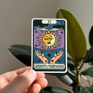 May include: A tarot card with a purple background and a sun and moon design. The card is labelled "XI" and "GRUMPY/SUNSHINE".