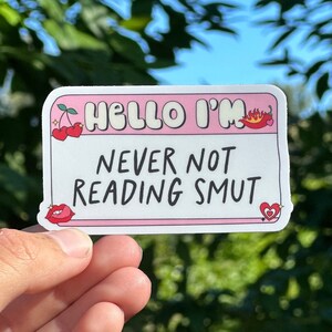 Puede incluir: Pegatina rosa y blanca con el texto "Hello I'm Never Not Reading Smut" con una cereza, un corazón y una cola de diablo.