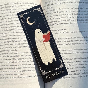 May include: A black bookmark with a white ghost reading a book. The bookmark has a crescent moon and the words "The Reader" on it.