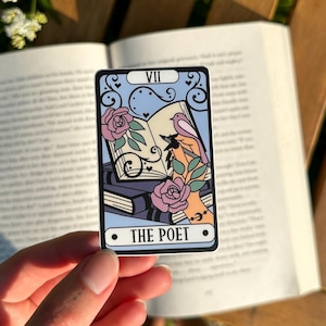May include: A tarot card with a black border, depicting a blue background with a hand holding a pen, roses, and books. The card reads "THE POET" at the bottom. A glass with a wooden lid and a metal straw is visible.