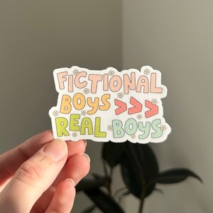 K&ouml;nnte beinhalten: Ein Aufkleber mit dem Text "Fictional Boys >>> Real Boys" in einem Regenbogen-Farbschema mit kleinen Blumenakzenten.