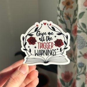 Peut inclure: Un autocollant blanc avec un motif de livre ouvert rouge. Le livre a une rose rouge de chaque côté et le texte "Give me all the trigger warnings" en noir.