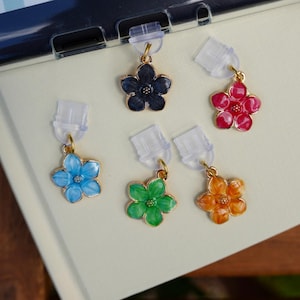 May include: A selection of flower-shaped earrings in blue, green, orange, red, and black. The earrings are suspended from clear plastic earring cards. A notebook with a dragon sticker and other stickers is in the background.