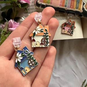 Peut inclure: Boucles d'oreilles en émail en forme de maison, bleues, oranges et roses, avec des détails floraux et de champignons. Une épingle avec une grenouille et un champignon, une coque de téléphone avec le texte "written by women" et un livre sont également visibles.
