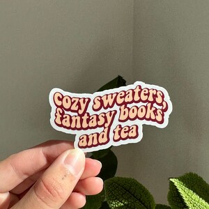 K&ouml;nnte beinhalten: Ein wei&szlig;er Aufkleber mit rotem Text, der "Cozy sweaters, fantasy books, and tea" lautet.
