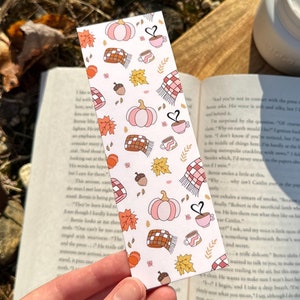 May include: A pink and orange bookmark with an autumn theme. It features pumpkins, leaves, acorns, coffee cups, and a scarf. The bookmark has a white background and is decorated with a heart.