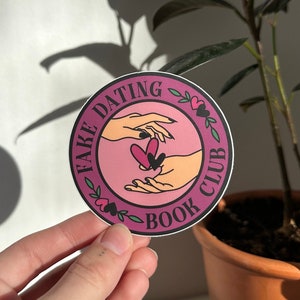 May include: A pink and white round sticker with the text "Fake Dating Book Club" and two hands holding a heart.