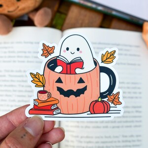 Op de afbeelding: Een Halloween-sticker met een cartoongeest die een boek leest in een pompoenmok. De sticker bevat herfstbladeren, een kleine pompoen en een stapel boeken. Het kleurenpalet omvat oranje, wit, zwart en rood.