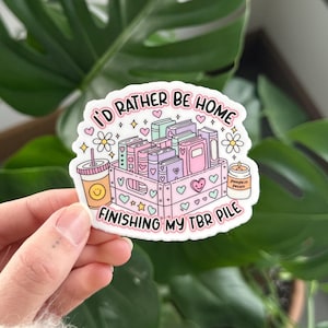 May include: A pink and white sticker with a cartoon illustration of a crate full of books, a coffee cup, and a candle. The text on the sticker reads "I'd rather be home finishing my TBR pile."