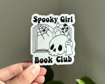 May include: A white sticker with a black line drawing of a skull, a book, a spider web, and a candle. The text "Spooky Girl Book Club" is written in black.