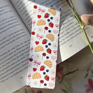 Pastel bookmark with a repeating pattern of croissants, strawberries, blueberries, flowers, bows, and hearts placed in front of an open book with soft floral bedding and dried flowers in the background