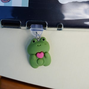 Op de afbeelding: Een groene kikkerhanger met een roze hart, hangend aan een doorzichtige plastic clip. De afbeelding toont ook stickers van een zeepaardje, zeester en een draak op een boek. De tekst op de afbeelding luidt: D230, 2FA CAN ICES-3(B), RG AMAZON.COM SERV, 73-5698 INPUT 5.0V 1.5A.