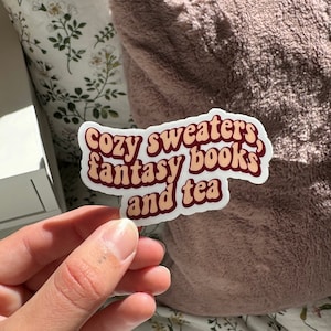 K&ouml;nnte beinhalten: Ein Aufkleber mit dem Text "Cozy sweaters, fantasy books and tea" in einer Retro-Schriftart mit braunen und orangen Farben.
