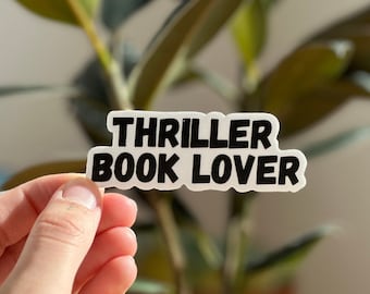 May include: White sticker with black text that reads "Thriller Book Lover". The sticker is being held in a hand with a potted plant in the background.