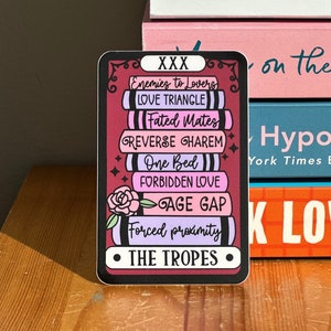 Puede incluir: Una pegatina con borde negro y fondo rosa y morado. La pegatina presenta una pila de libros con el texto "XXX" en la parte superior y "THE TROPES" en la parte inferior. Los libros están etiquetados con tropos románticos comunes, que incluyen "Enemies to Lovers", "Love Triangle", "Fated Mates", "Reverse Harem", "One Bed", "Forbidden Love", "Age Gap" y "Forced Proximity".