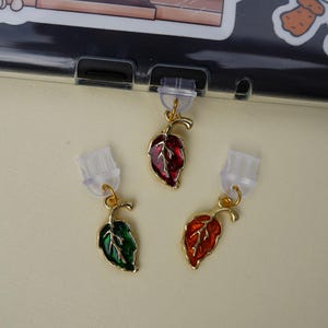 May include: Three leaf-shaped earrings in red, green, and orange enamel with gold-coloured metal accents. The earrings are displayed against a light background, with stickers of a bookstore and a duck in a sweater.