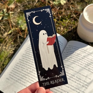 May include: A black bookmark featuring a white ghost reading a red book. The bookmark has a crescent moon and stars in the background. The text "THE READER" is printed on the bookmark.