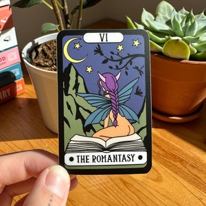 May include: A tarot card with a black border and a purple fairy with wings sitting on a book. The card is labelled "VI" and "THE ROMANTASY".
