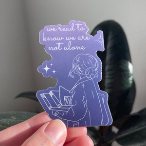 May include: A purple and blue sticker with a line drawing of a person reading a book. The sticker has the text "we read to know we are not alone" and stars.