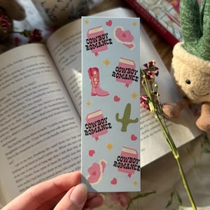May include: A blue bookmark with pink and white illustrations of cowboy boots, books, cacti, and hearts. The text "Cowboy Romance" is repeated on each book illustration.