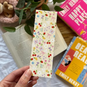 May include: A cream-coloured bookmark featuring a pattern of cherries, strawberries, butterflies, and flowers. The bookmark is held in a hand, with two books and a potted plant in the background.