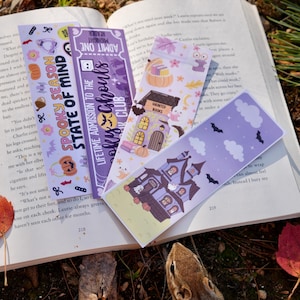 Può includere: Segnalibri a tema Halloween con case infestate, pipistrelli e zucche. Sono appoggiati su un libro aperto, con uno scoiattolo in primo piano.
