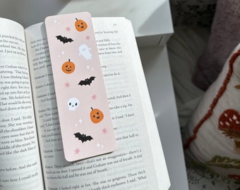 May include: A pink and white Halloween-themed bookmark with illustrations of pumpkins, bats, ghosts, and a skull. The bookmark has a pink background with white stars and black outlines. The text on the bookmark reads "Lane Three seconds later that's not an exaggeration there's a knock on my door. I opened it and Graham was out of breath. A tiny man draws the hall and he was out of breath."