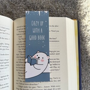May include: A blue and white bookmark featuring a polar bear reading a book. The bookmark has the text "Cozy Up With A Good Book" on it.