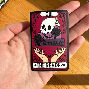 May include: A tarot card with a black border and a red background. The card features a skull with a candle on top of it, and the words "Dark Romance" written below. The card is labeled "XIII" and "THE READER" in black text.