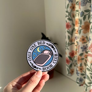May include: A round sticker with a blue background and white border. The sticker features a bed with a pink pillow and a crescent moon with stars. The text "Only One Bed Book Club" is written in black around the bed.