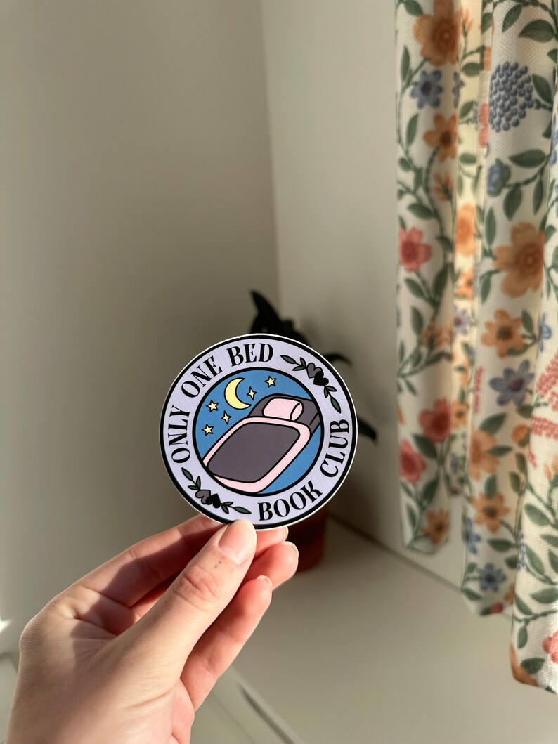 May include: A round sticker with a blue background and white border. The sticker features a bed with a pink pillow and a crescent moon with stars. The text "Only One Bed Book Club" is written in black around the bed.