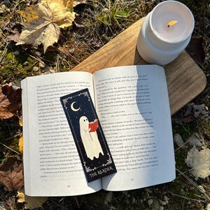May include: A black bookmark with a white ghost reading a book. The bookmark has the words "The Reader" on it. The bookmark is on top of an open book with text about a ghost.