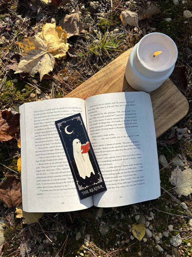 May include: A black bookmark with a white ghost reading a book. The bookmark has the words "The Reader" on it. The bookmark is on top of an open book with text about a ghost.