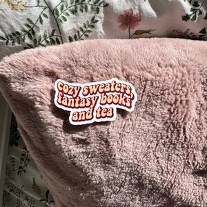 K&ouml;nnte beinhalten: Eine rosa flauschige Decke mit einem wei&szlig;en Aufkleber, auf dem "Cozy sweaters, fantasy books, and tea" steht.