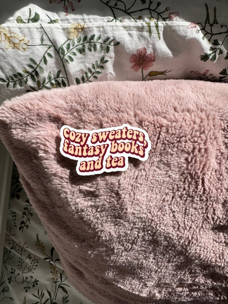 K&ouml;nnte beinhalten: Eine rosa flauschige Decke mit einem wei&szlig;en Aufkleber, auf dem "Cozy sweaters, fantasy books, and tea" steht.