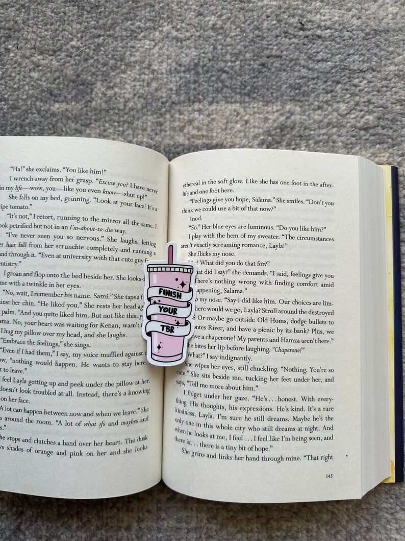 K&ouml;nnte beinhalten: Nahaufnahme eines rosa und wei&szlig;en Buches mit den Worten "Finish Your TBR" auf einem rosa Lesezeichen. Das Buch ist auf einer Seite mit Text &uuml;ber die Gef&uuml;hle einer jungen Frau f&uuml;r einen Mann aufgeschlagen. Der Text lautet: "Feelings give you hope, Salama."