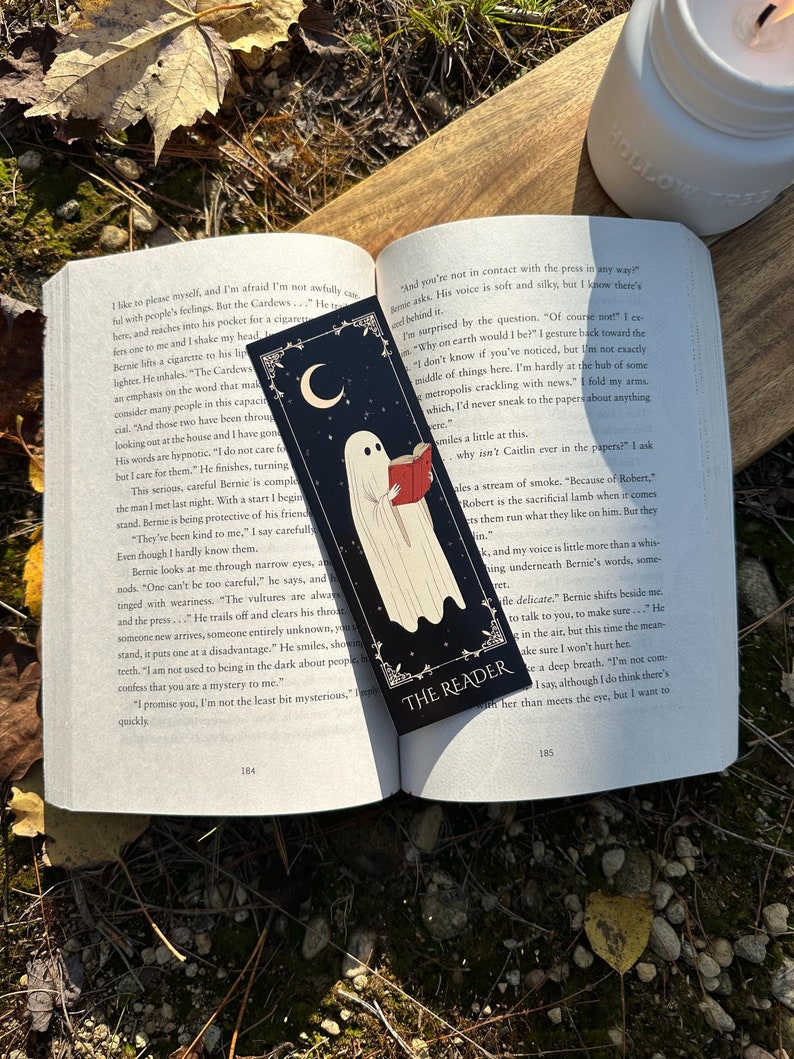 May include: A black bookmark with a white ghost reading a book. The bookmark has a crescent moon and the words "The Reader" on it.