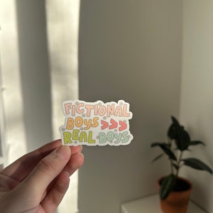 K&ouml;nnte beinhalten: Ein Aufkleber mit dem Text "Fictional Boys >>> Real Boys" in einem Regenbogen-Farbverlauf. Der Aufkleber ist mit kleinen wei&szlig;en Blumen verziert.