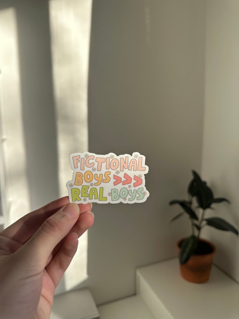 K&ouml;nnte beinhalten: Ein Aufkleber mit dem Text "Fictional Boys >>> Real Boys" in einem Regenbogen-Farbverlauf. Der Aufkleber ist mit kleinen wei&szlig;en Blumen verziert.