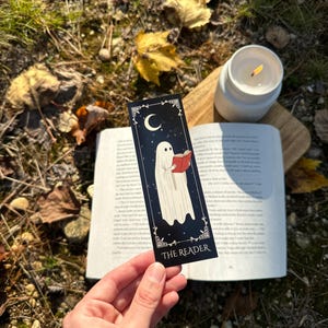 May include: A black bookmark with a white ghost reading a red book. The bookmark has a crescent moon and stars in the background. The text "THE READER" is at the bottom of the bookmark.