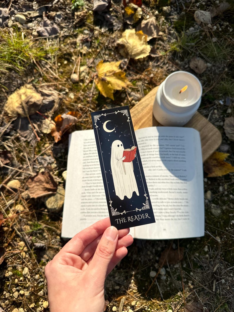 May include: A black bookmark with a white ghost reading a red book. The bookmark has a crescent moon and stars in the background. The text "THE READER" is at the bottom of the bookmark.