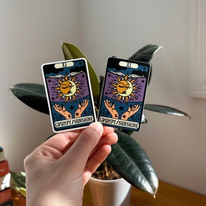 May include: Two stickers featuring a sun and moon with a face, a book, and hands. The stickers are on a white background with the text "XI GRUMPY/SUNSHINE".