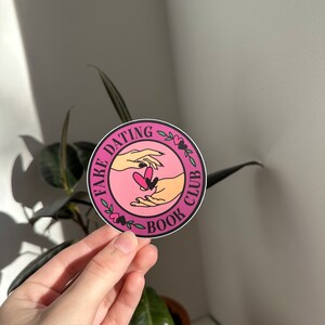 May include: A pink and black round sticker with the text "Fake Dating Book Club" and a graphic of two hands holding a heart.