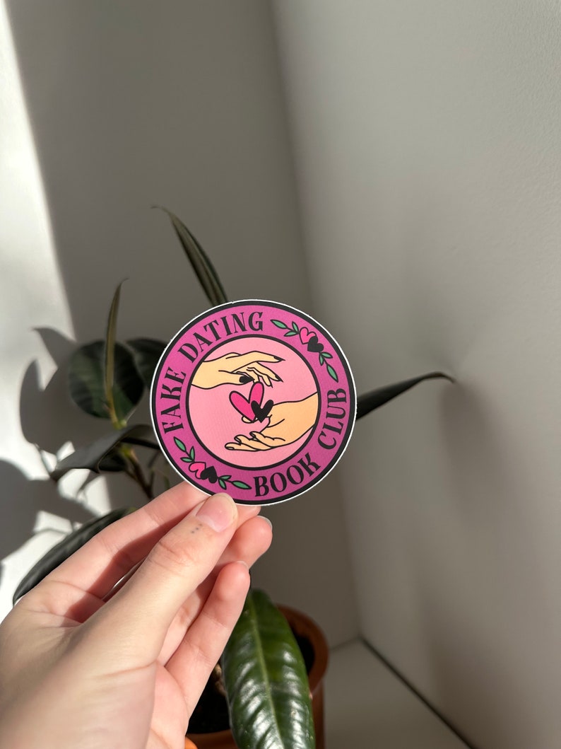 May include: A pink and black round sticker with the text "Fake Dating Book Club" and a graphic of two hands holding a heart.