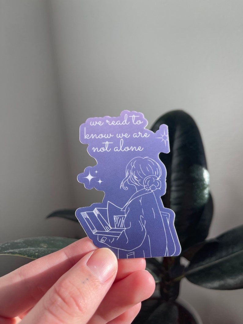 May include: A purple and blue sticker with a line drawing of a person reading a book. The sticker has the text "we read to know we are not alone" and stars.