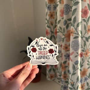 May include: A white sticker with a red and black design. The sticker features a book with a floral wreath and the text "Give me all the trigger warnings".