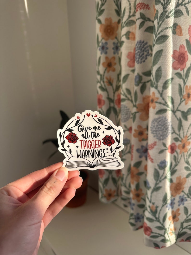 May include: A white sticker with a red and black design. The sticker features a book with a floral wreath and the text "Give me all the trigger warnings".