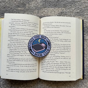 May include: A close-up of a book with the title "Only One Bed Book Club" printed on a blue and white enamel pin. The pin features a bed with a single person sleeping in it. The book is open to a page with text about a young woman's feelings for a man.
