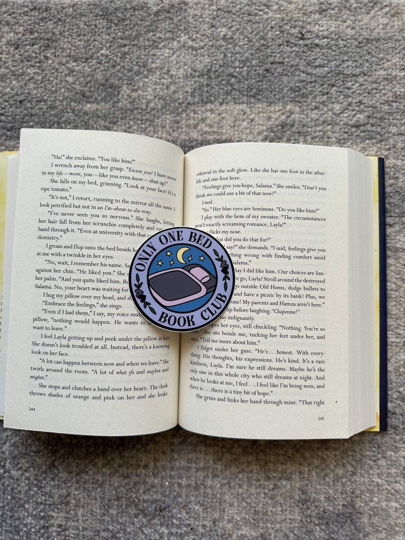 May include: A close-up of a book with the title "Only One Bed Book Club" printed on a blue and white enamel pin. The pin features a bed with a single person sleeping in it. The book is open to a page with text about a young woman's feelings for a man.